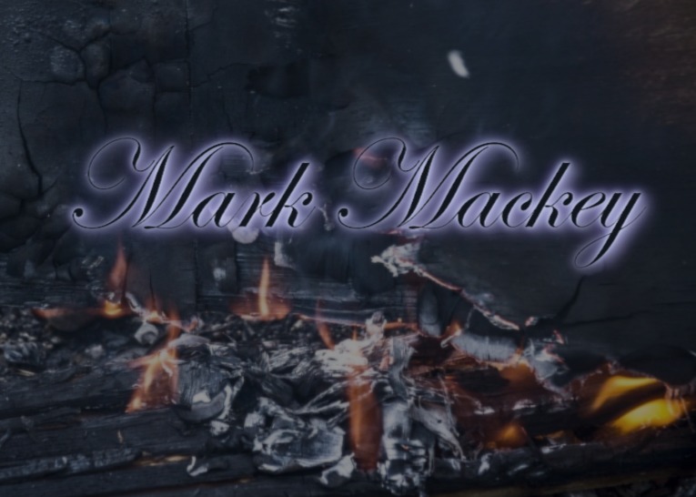 The name 'Mark Mackey' displayed in an elegant font against a background of smoldering ashes and embers.
